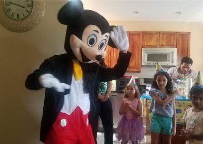 A costumed Mickey Mouse figure greets a group of children at a birthday party in a kitchen setting.