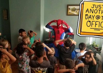 A person in a Spider-Man costume stands with a group of children who all have their hands on their heads in an indoor room.