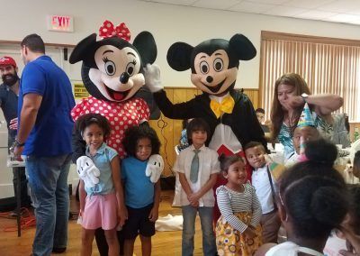 Mickey and Minnie Mouse characters waving and holding hands in front of a stone-textured wall.