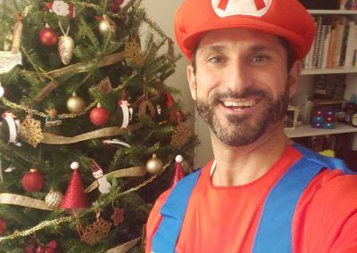 A smiling person wearing a Mario costume and hat, standing in front of a decorated Christmas tree.