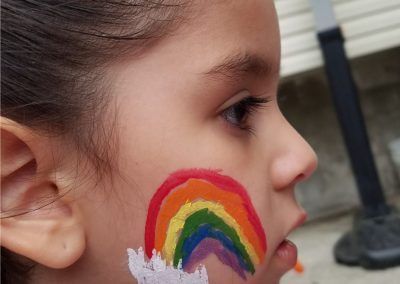 A child's cheek is painted with a vibrant, colorful rainbow featuring red, orange, yellow, green, blue, and purple hues.