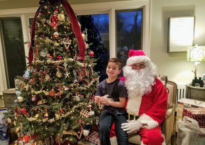 A smiling child sits on Santa’s lap next to a decorated Christmas tree in a living room.