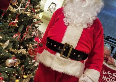 Santa Claus standing in front of a decorated Christmas tree with a person waving in the background.
