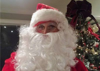 A person in a Santa Claus costume with a white beard and hat, standing in front of a decorated Christmas tree.