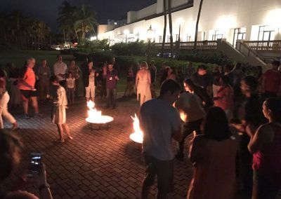 A group of people gathers outdoors at night around two small fire pits on a brick patio in front of a building.