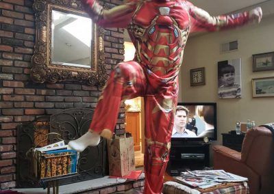 A person wears an Iron Man costume in a living room, standing on one leg in front of a brick fireplace and television.