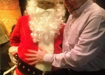 A person in a Santa Claus costume has their belly touched by another individual in a light-colored dress shirt.