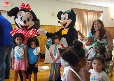 Mickey and Minnie Mouse mascots pose for photos with a group of children and an adult in a brightly lit indoor venue.