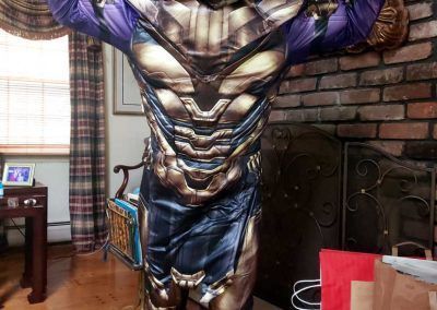 A person wearing a Thanos costume with detailed gold and purple armor, standing in a room with a brick fireplace.
