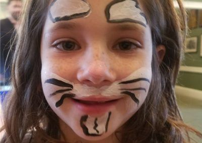 A child with cat face paint, featuring white patches over the eyes and chin, black whisker lines, and a light smile.