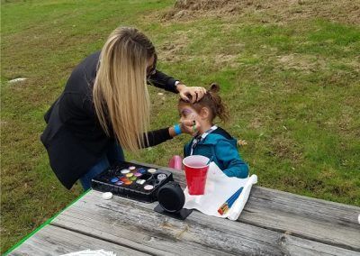 A person paints a child's face at an outdoor table with supplies and a red plastic cup.