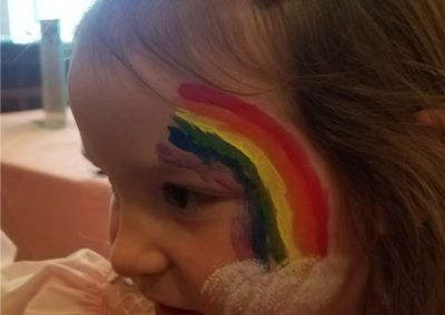 A side-profile view of a child with a vibrant, multicolored rainbow face painting extending across their temple.
