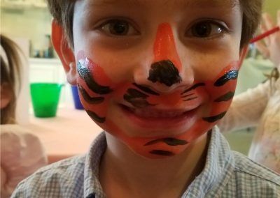 A close-up of a child wearing orange and black tiger face paint, smiling, with a blurry room in the background.