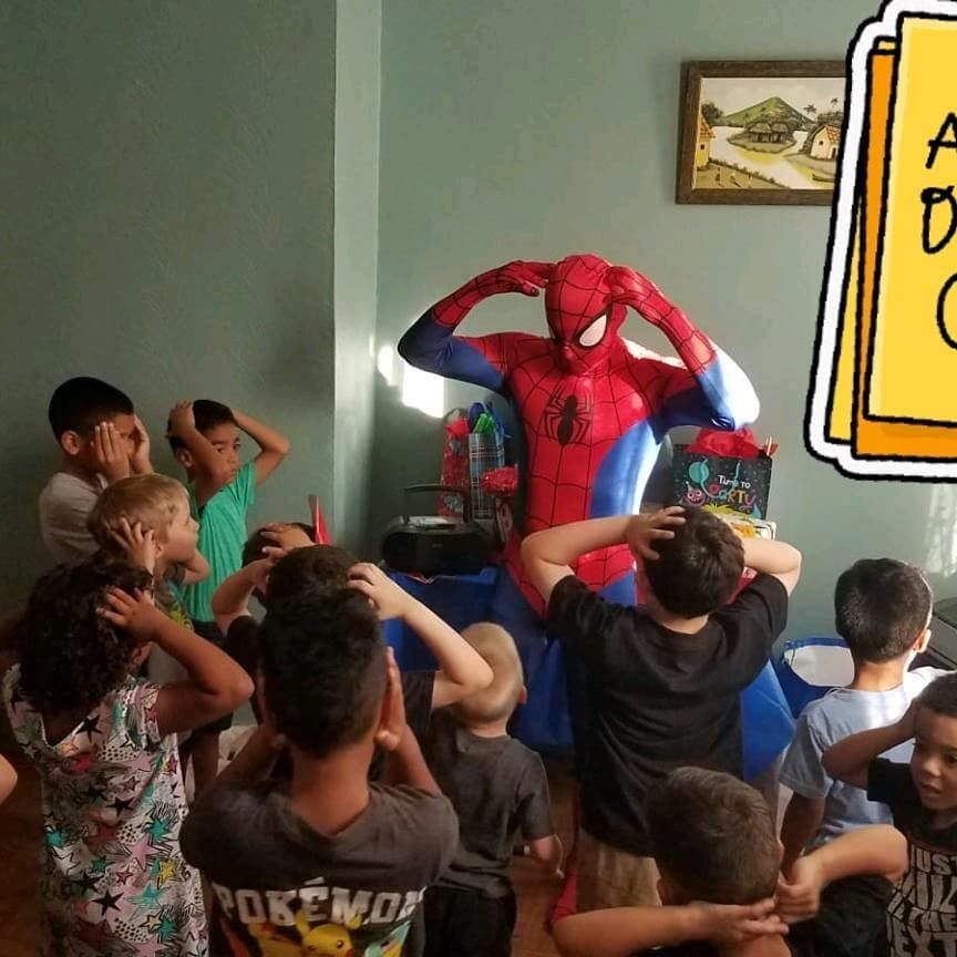A costumed Spider-Man mimics a group of children, all with hands on their heads, in a dimly lit indoor room.