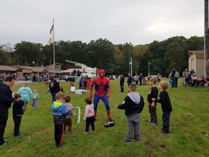 A person in a Spider-Man costume stands in a grassy field surrounded by children during an outdoor community event.