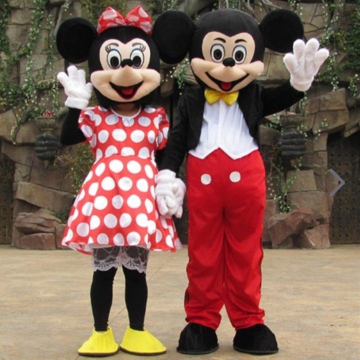 Mickey and Minnie Mouse mascots in classic outfits stand side-by-side, waving in front of a stone-textured wall.