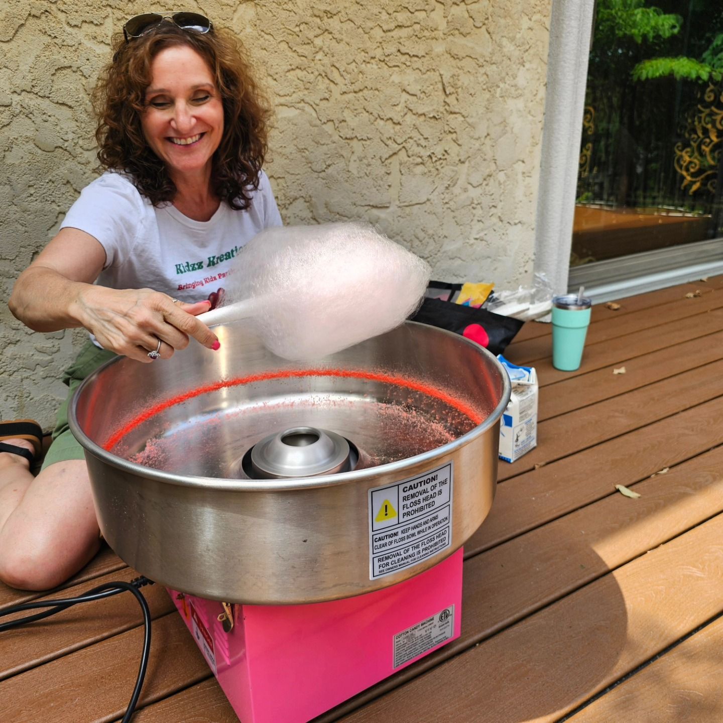 A smiling person uses a pink cotton candy machine on a wooden deck to spin a cloud of pink sugar onto a cone.