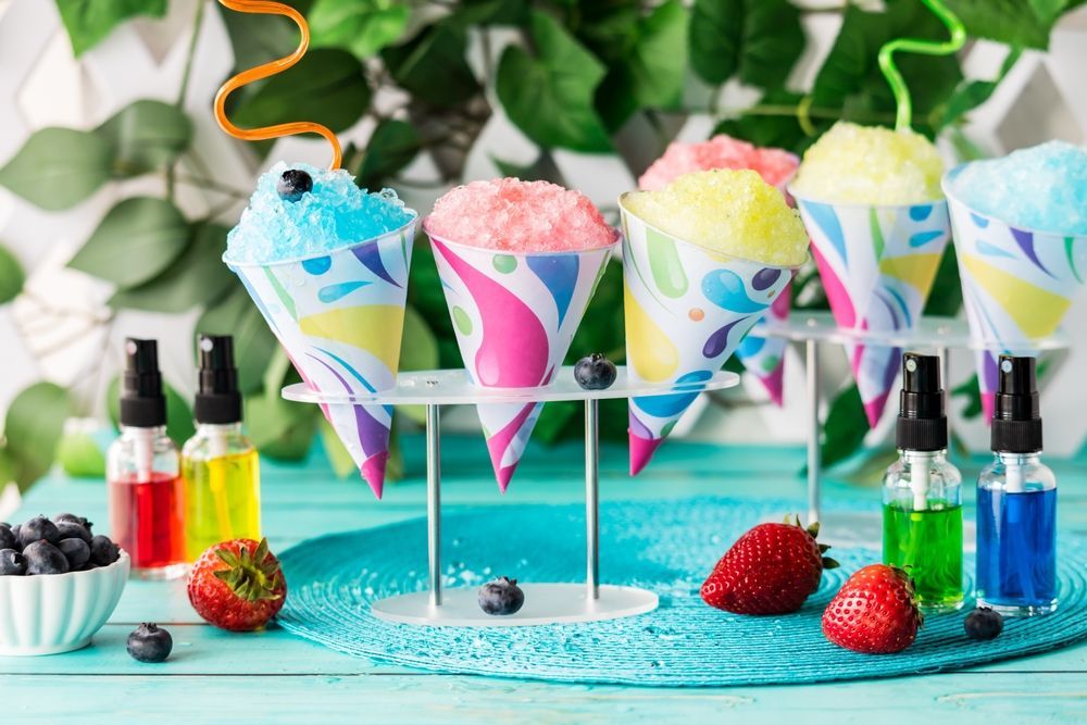 Five colorful snow cones in patterned paper cups on a stand, surrounded by fresh strawberries, blueberries, and syrups.
