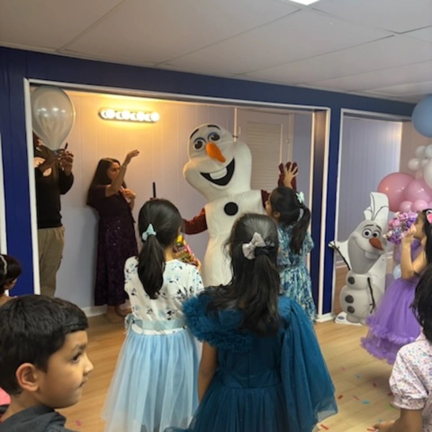 Children in party attire gather around a large Olaf character mascot in an indoor room decorated with balloons.