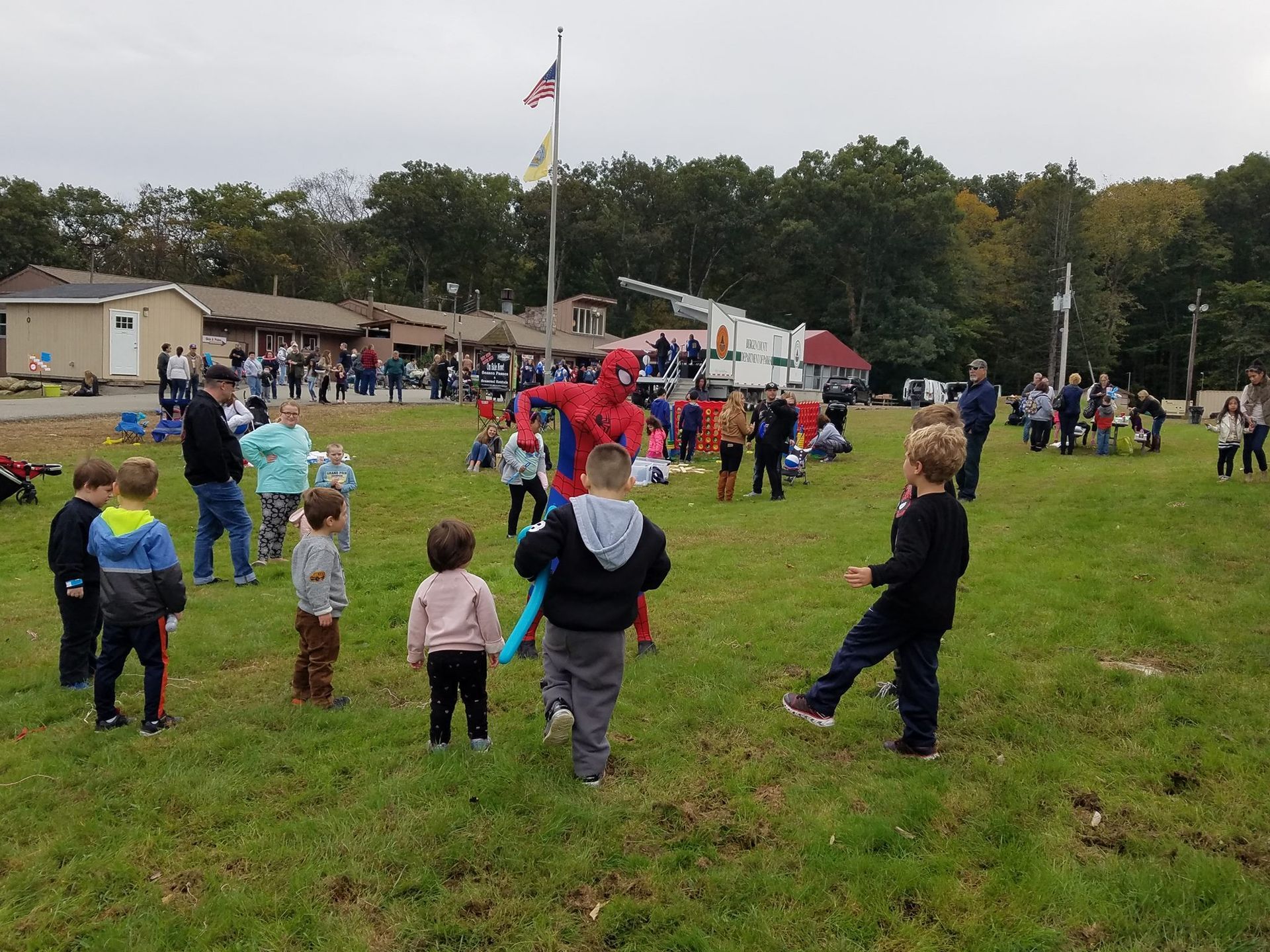 A person in a Spider-Man costume interacts with children in a grassy field during an outdoor event near a building.