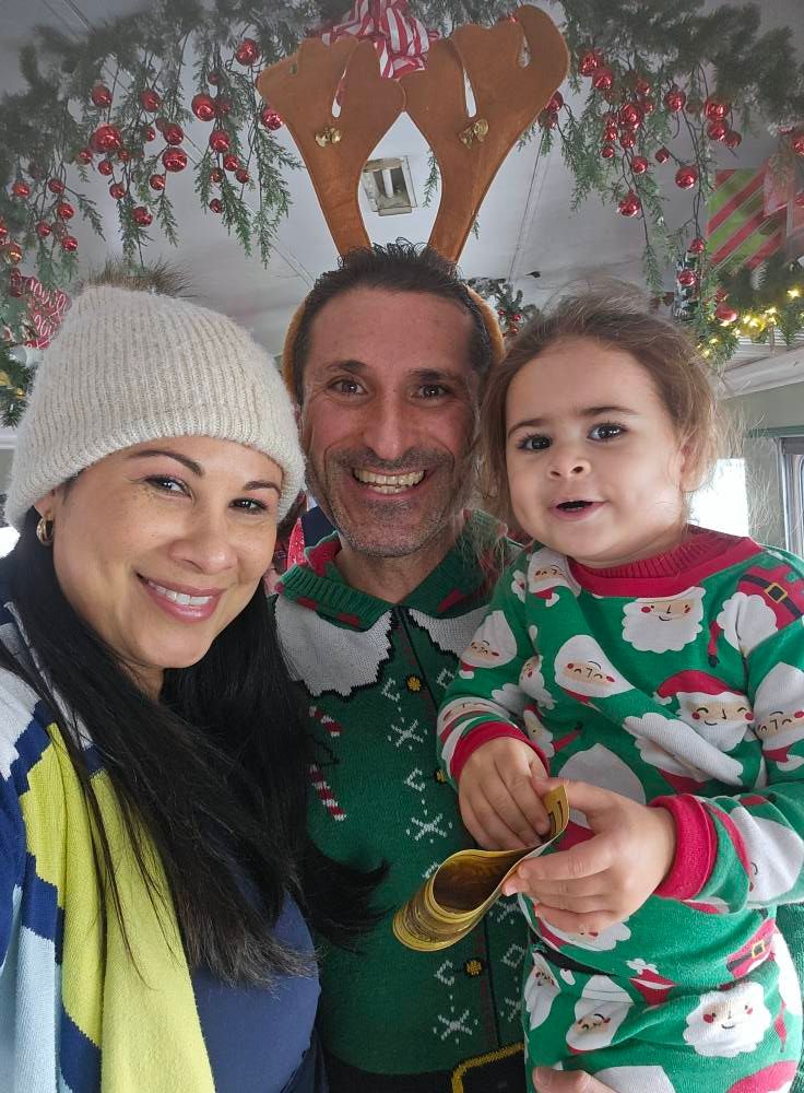 A person with reindeer antlers, a woman in a beanie, and a child in holiday pajamas smile together in a festive room.