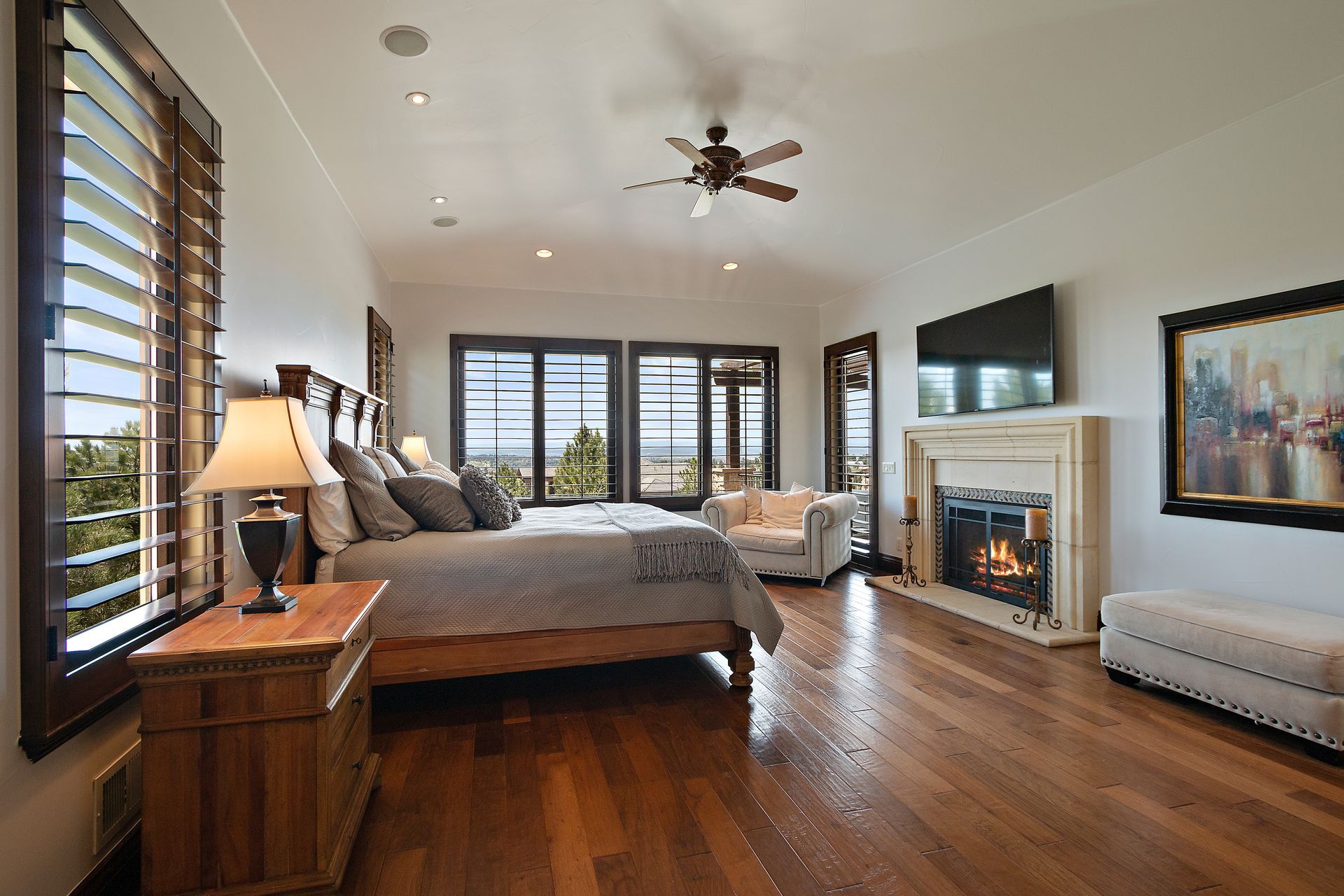 Spacious bedroom with wood floors, bed, fireplace, large windows overlooking view, and a ceiling fan.