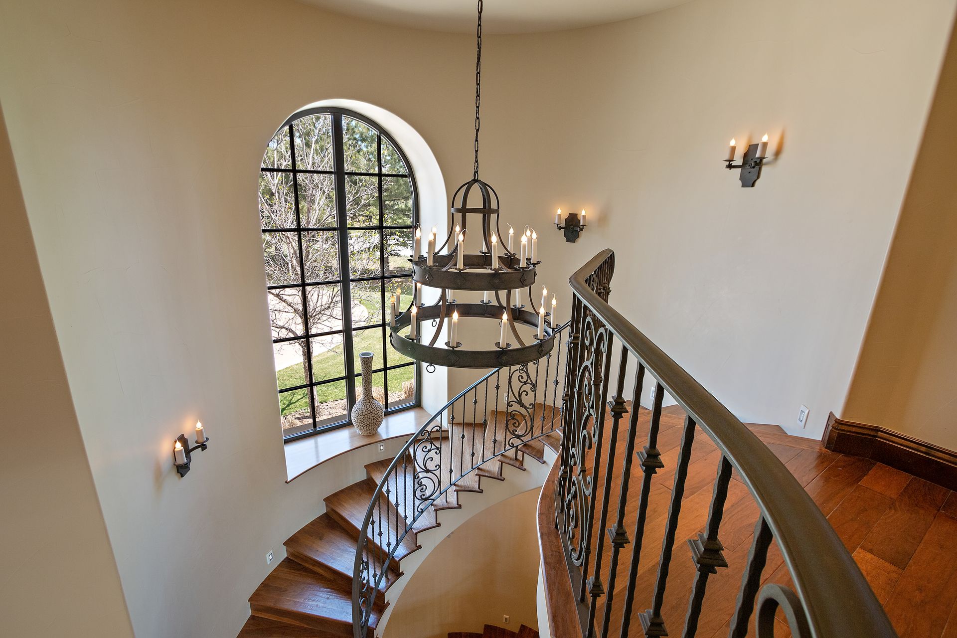 A curved staircase with iron railing and a chandelier, next to a large arched window.