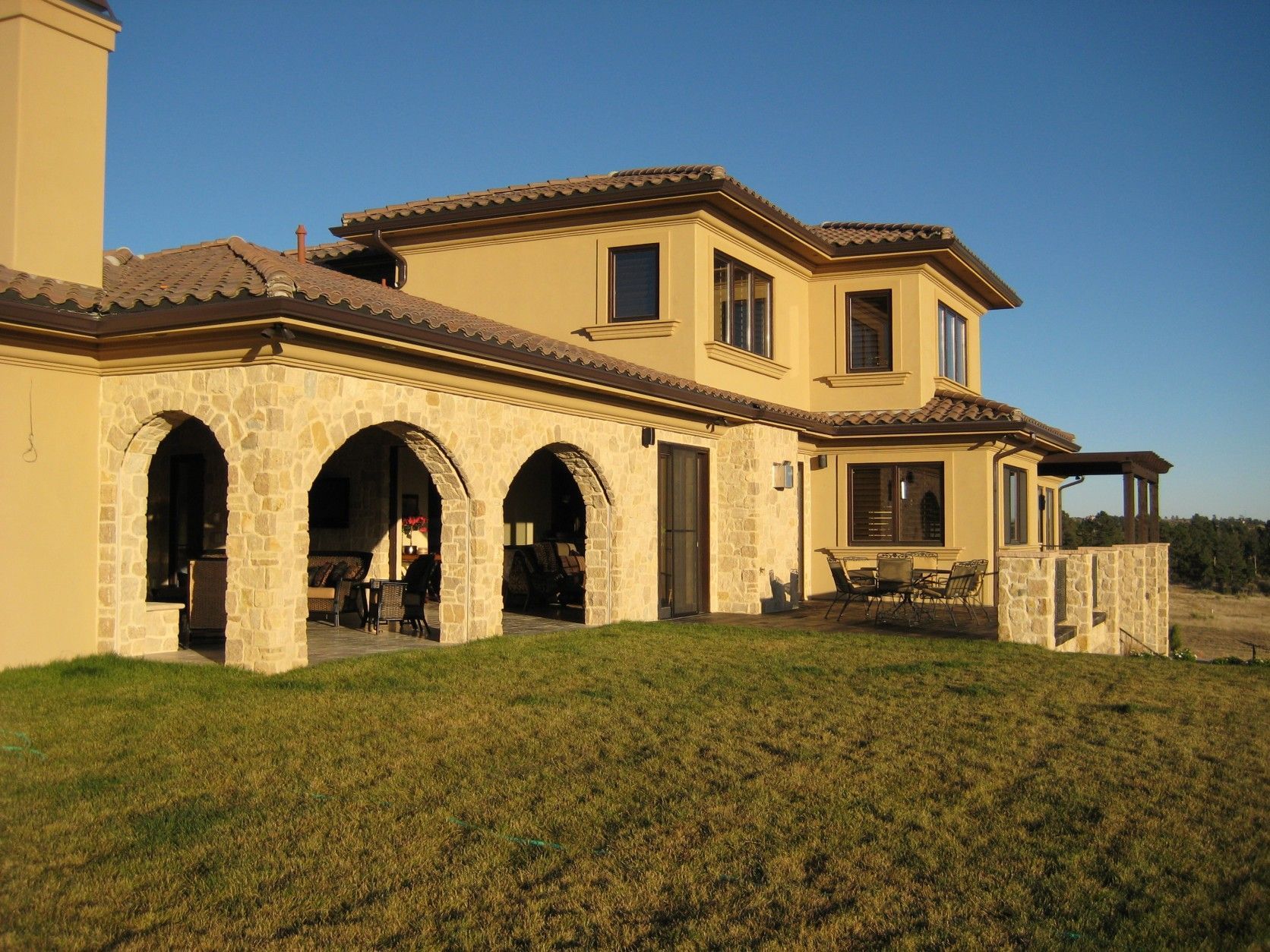 Tan stucco home with stone arches and a tiled roof on a grassy lawn under a clear blue sky.