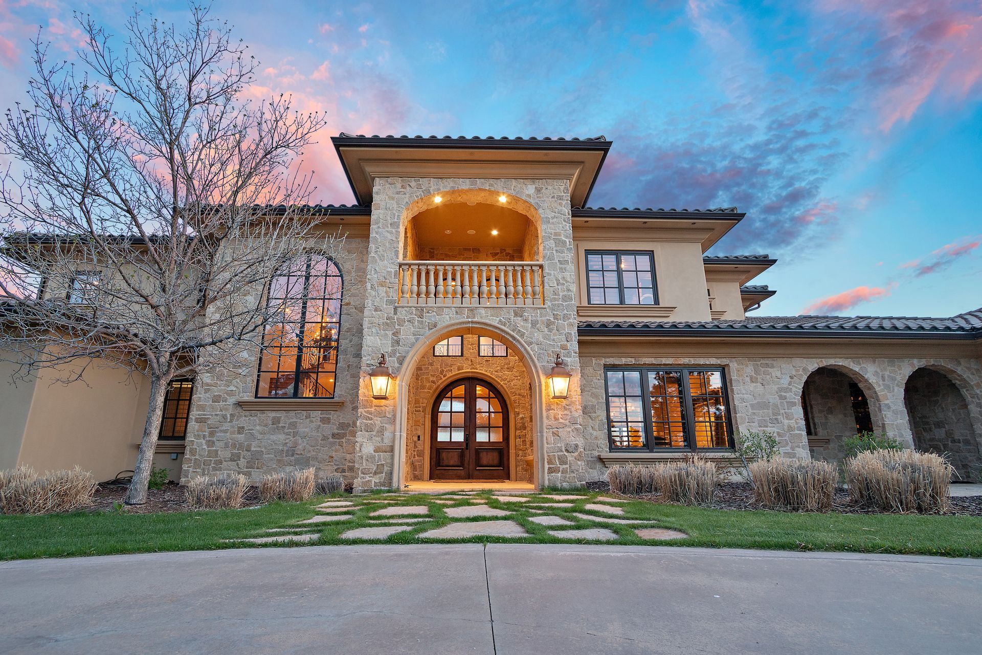Luxurious two-story home with stone facade, arched doorway, balcony, and sunset sky.