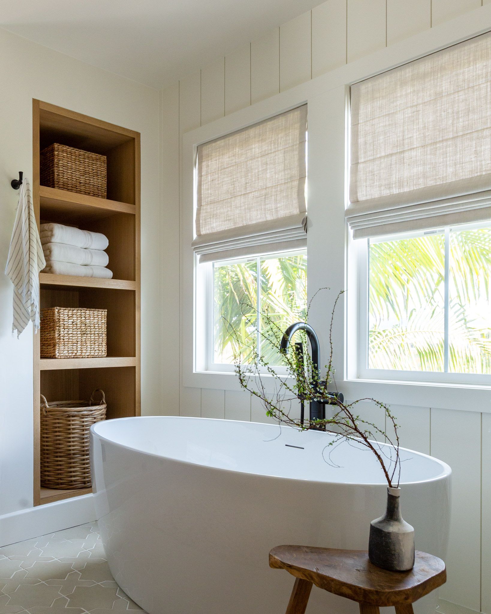 Cozy bathroom with a white tub, woven blinds, wooden shelves with baskets, and a small wooden stool.