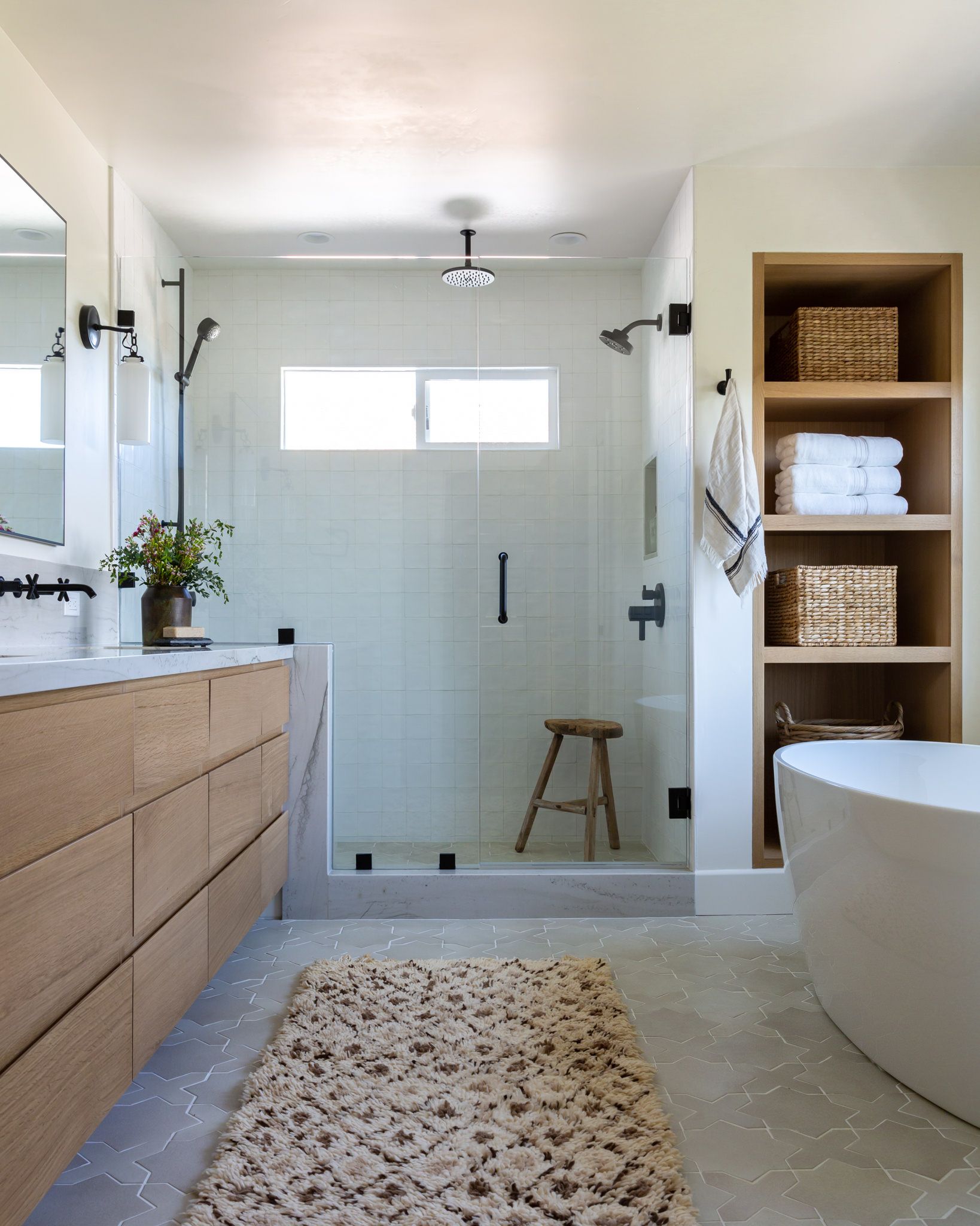 Modern bathroom with wood vanity, glass shower, and built-in shelving. Neutral tones, cozy rug, and a freestanding tub.