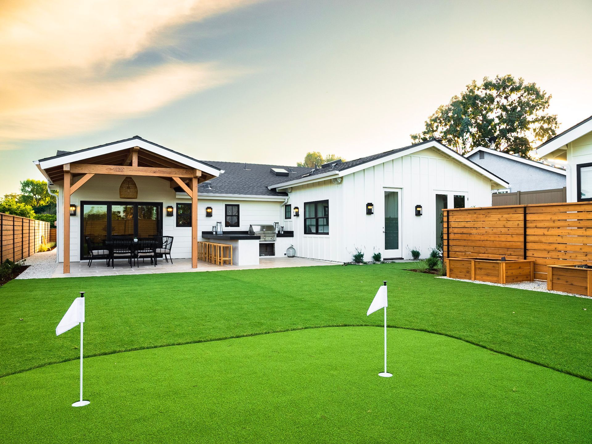Backyard with putting green, white house, wooden pergola and fence, green grass, sunny.