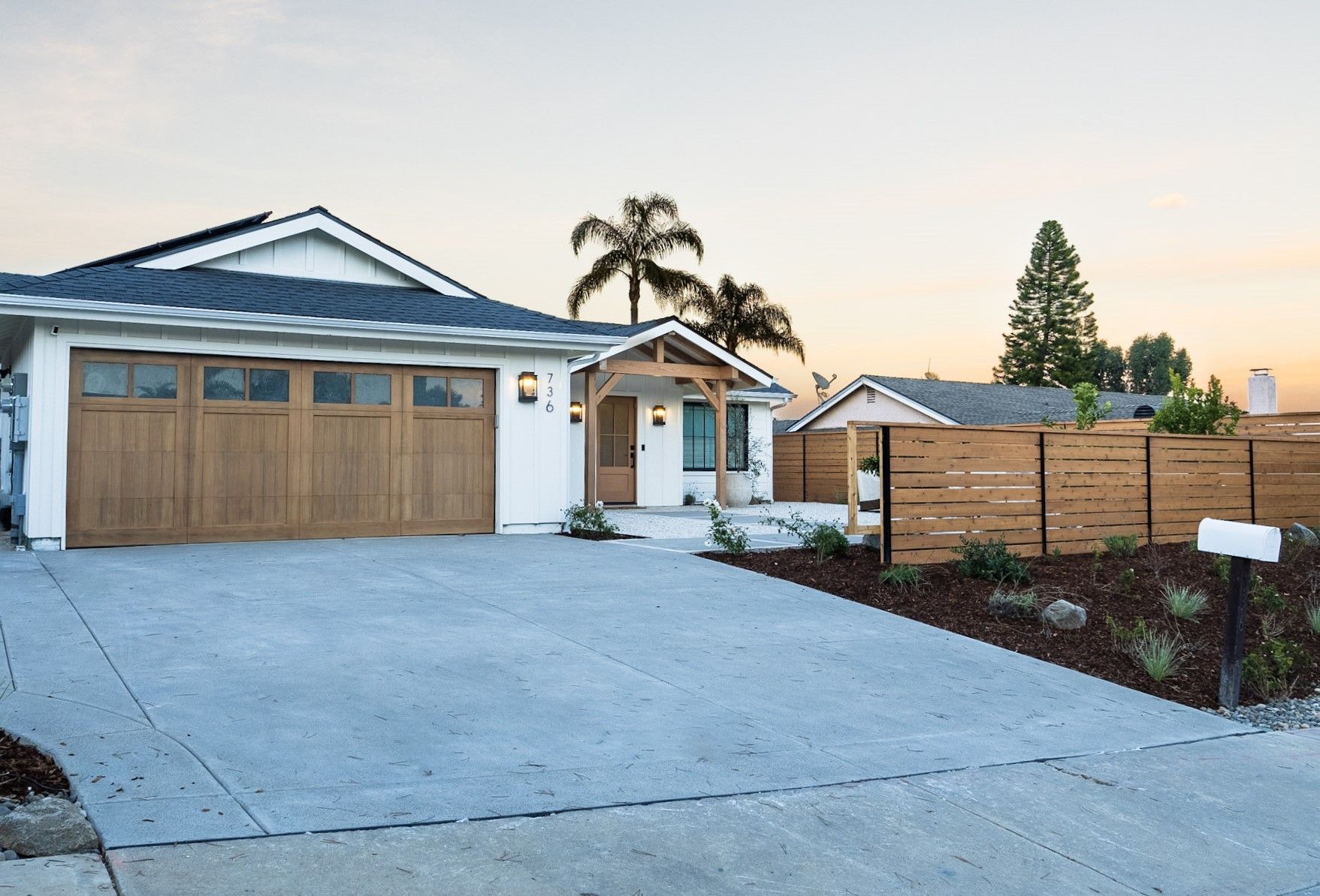 White house with wooden garage door, driveway, wooden fence, and palm trees.
