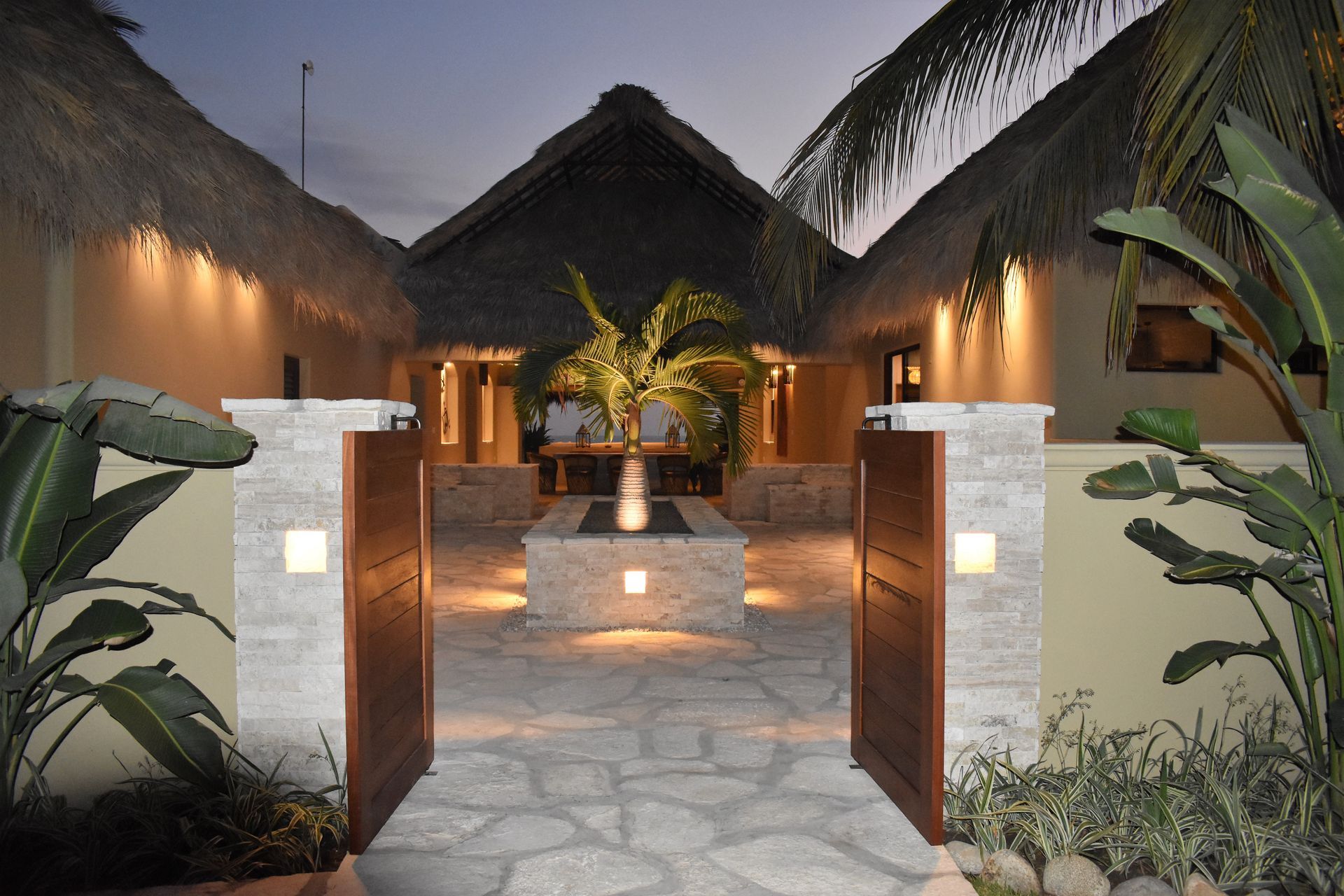 Open wooden gate leading to a tropical courtyard with thatched roof buildings and a lit palm tree.