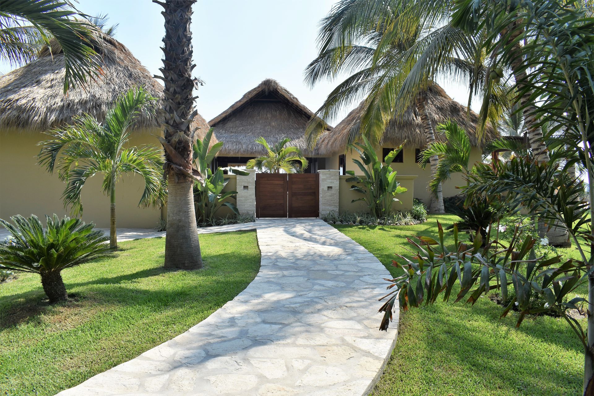 Stone path leads to a tan building with thatched roofs and wooden gates, surrounded by tropical plants.