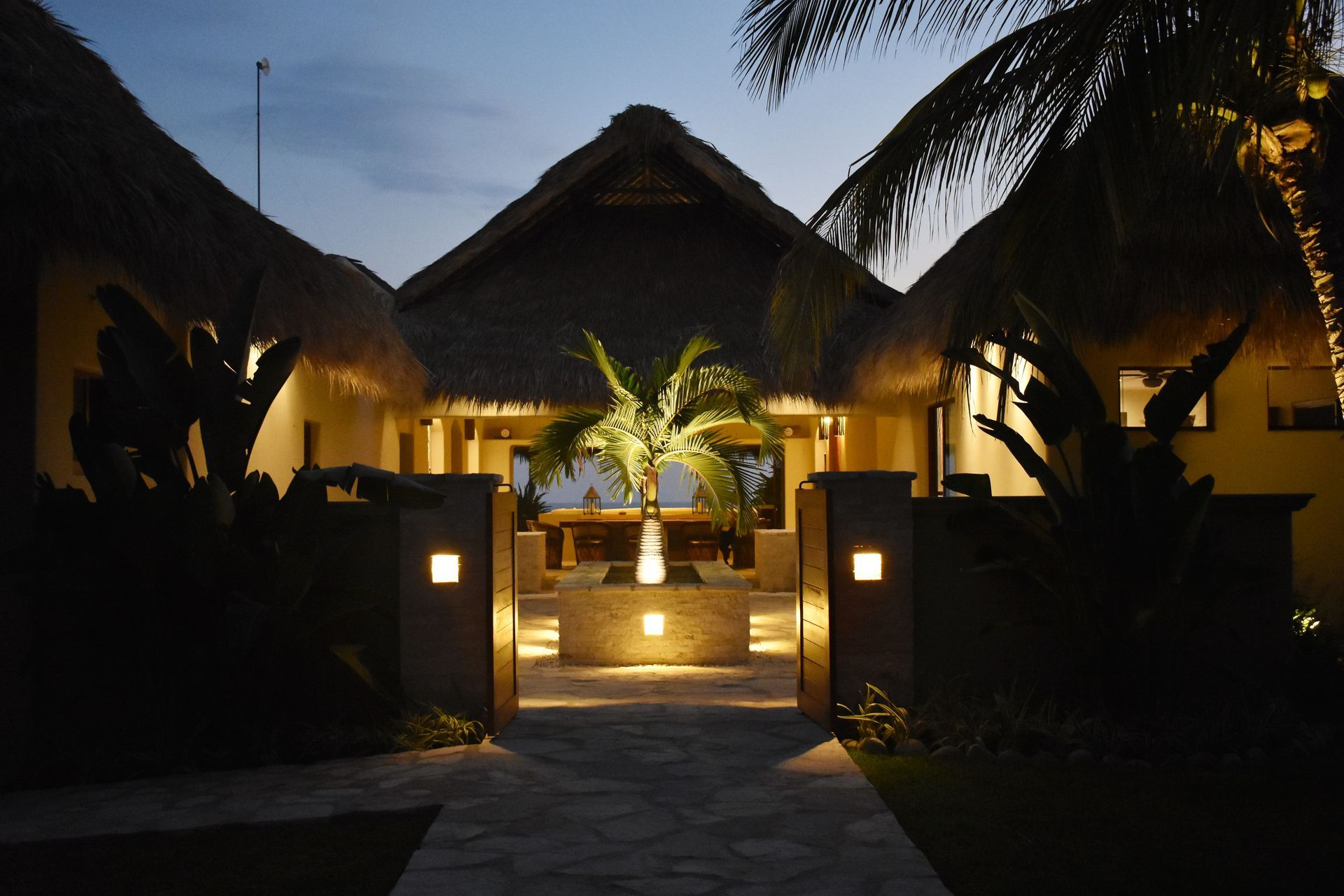Night view of a thatched-roof building with pathway lit by ground lights. Tropical foliage and palm trees.