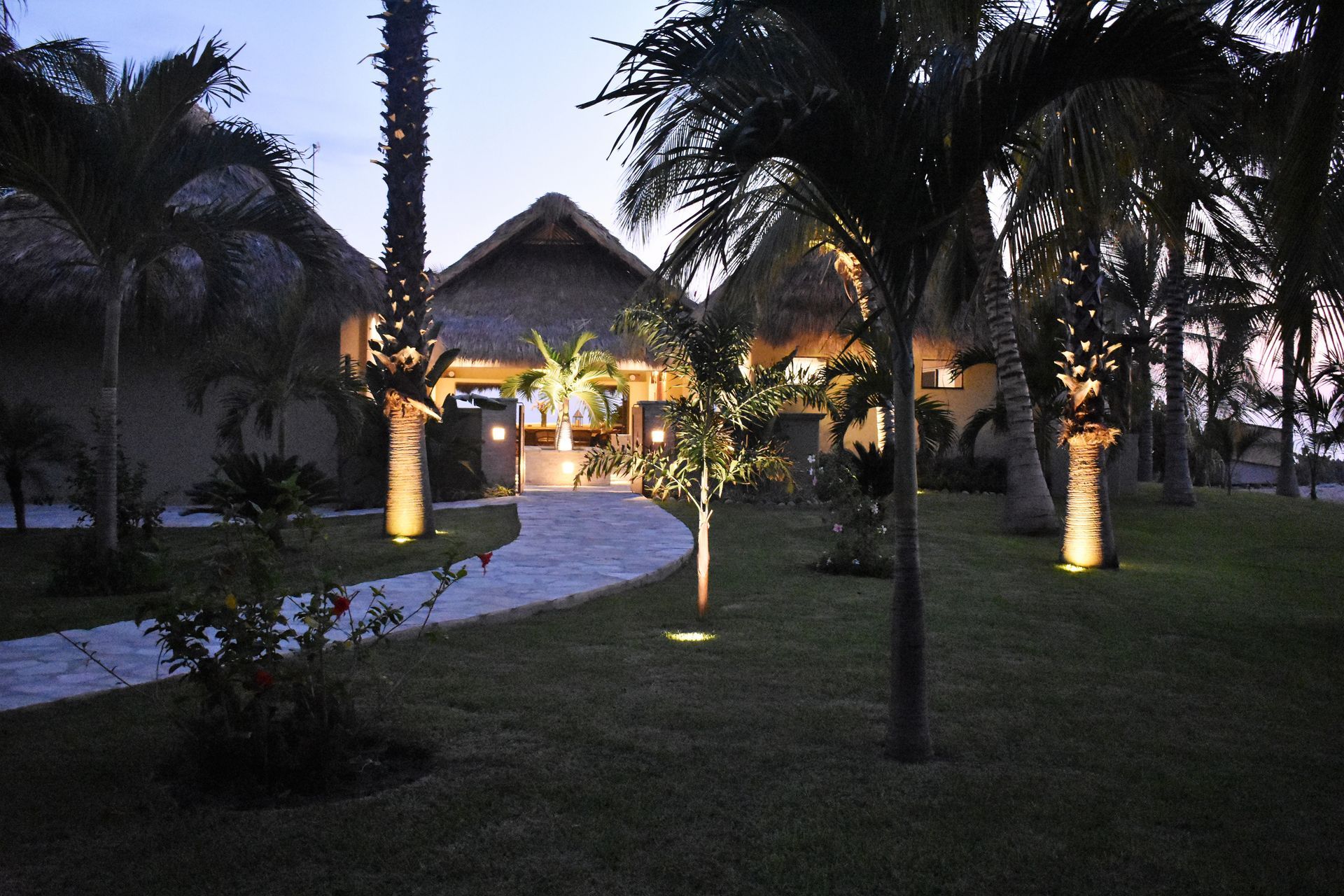Stone path leads to thatched-roof buildings with palm trees, illuminated at dusk.