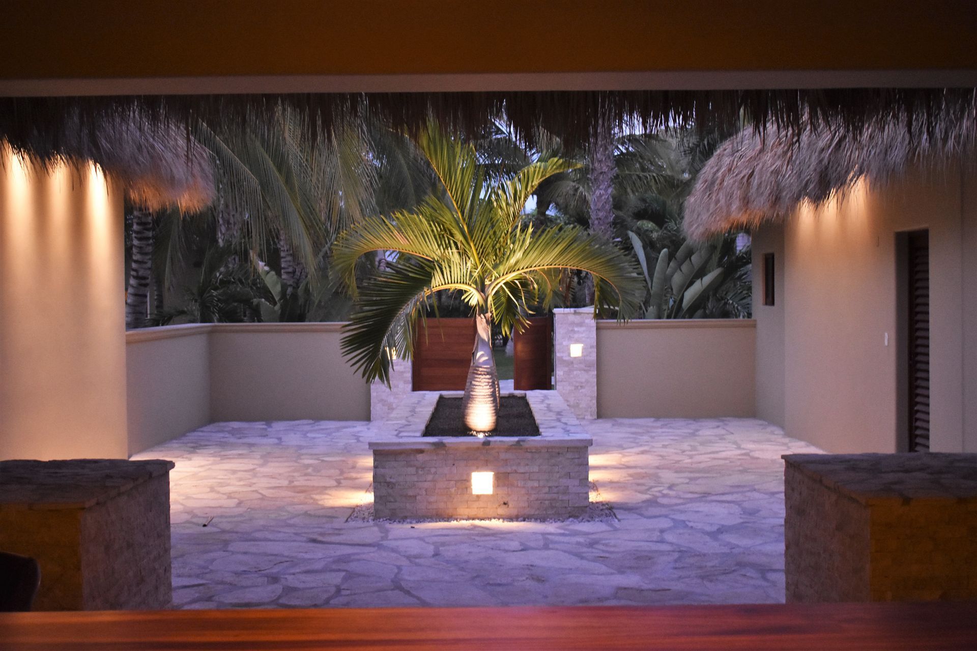 Tropical courtyard with palm tree, lit with warm lights, stone pavers, and thatched roof.