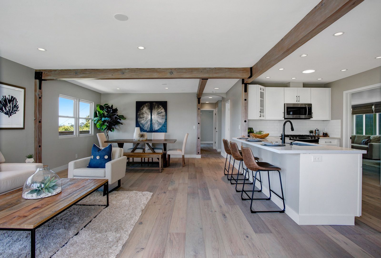 Open-concept kitchen and living room with light wood floors, white cabinets, and wooden beams.