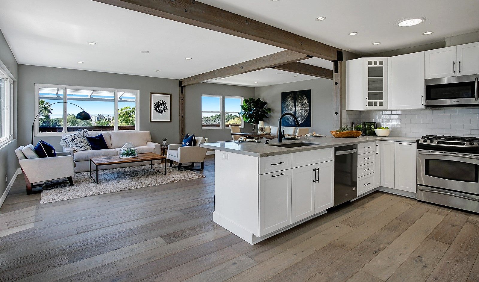 Open-concept kitchen and living room with white cabinets, wood floors, and exposed ceiling beams.