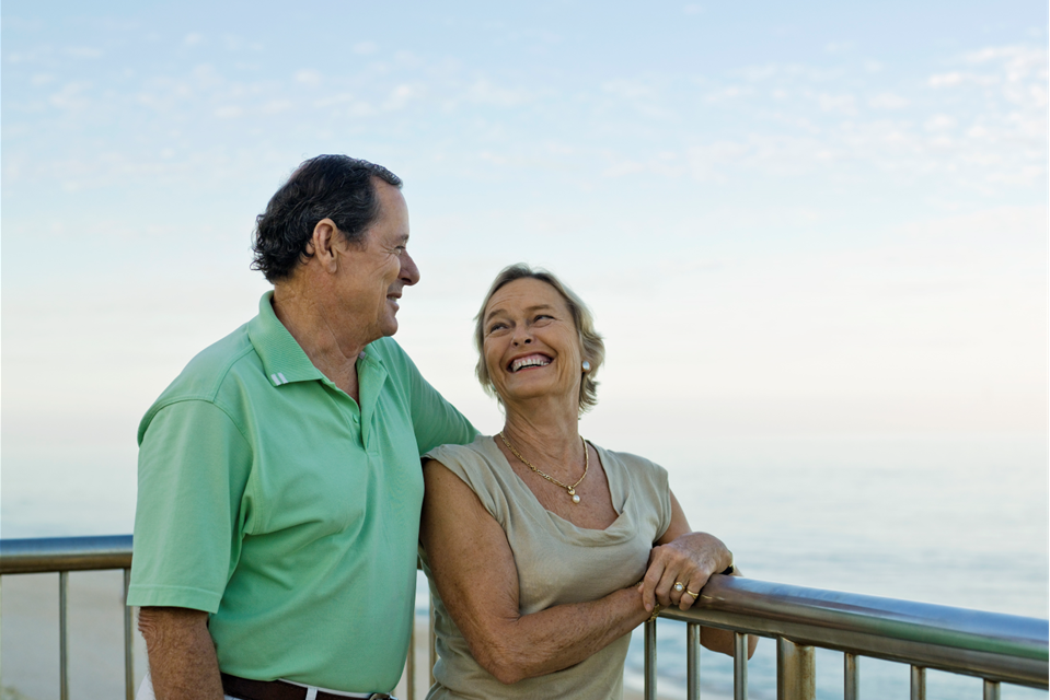 Smiling couple overlooking the ocean from a balcony; man in green shirt, woman in tan top.