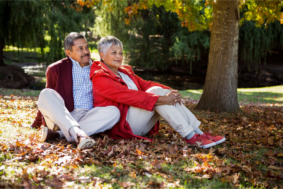 Couple sitting together under a tree on a bed of leaves; one in a red coat, the other in a maroon jacket.