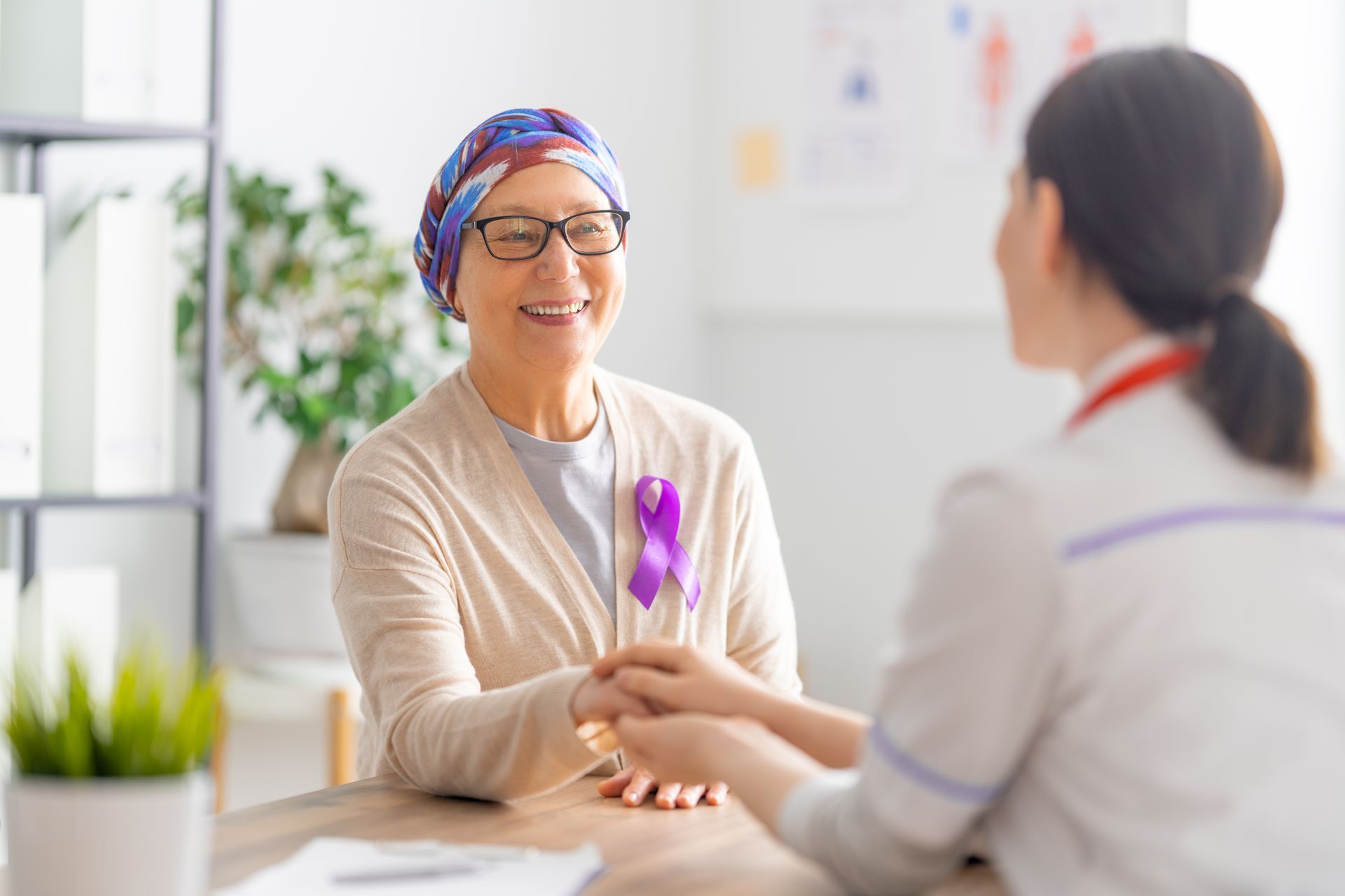 Woman with headscarf and purple ribbon smiles as a doctor holds her hand at a desk.