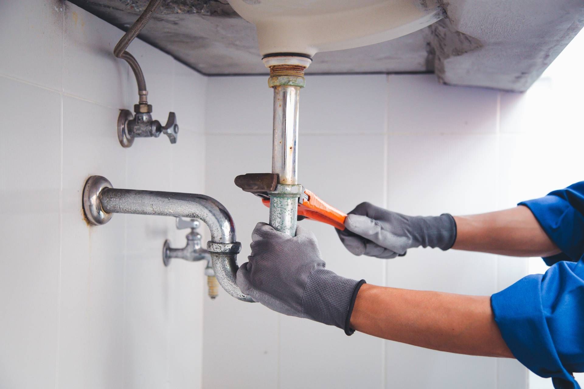 Plumber with grey gloves using a wrench to repair sink pipes in a bathroom.