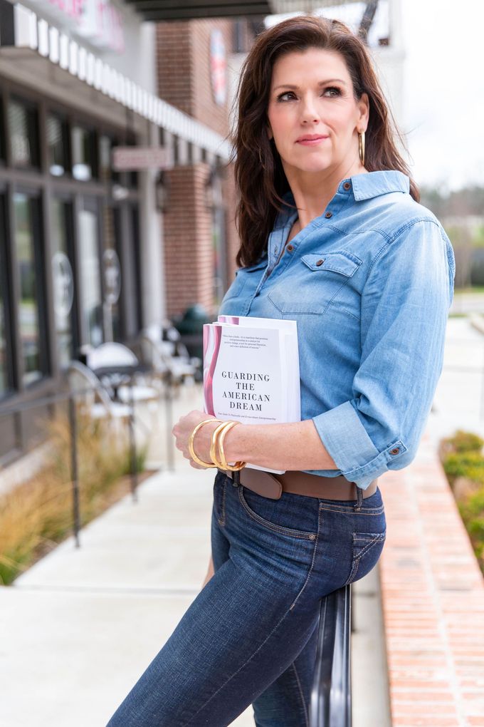 a woman in a denim shirt and jeans is standing on a sidewalk holding a book .