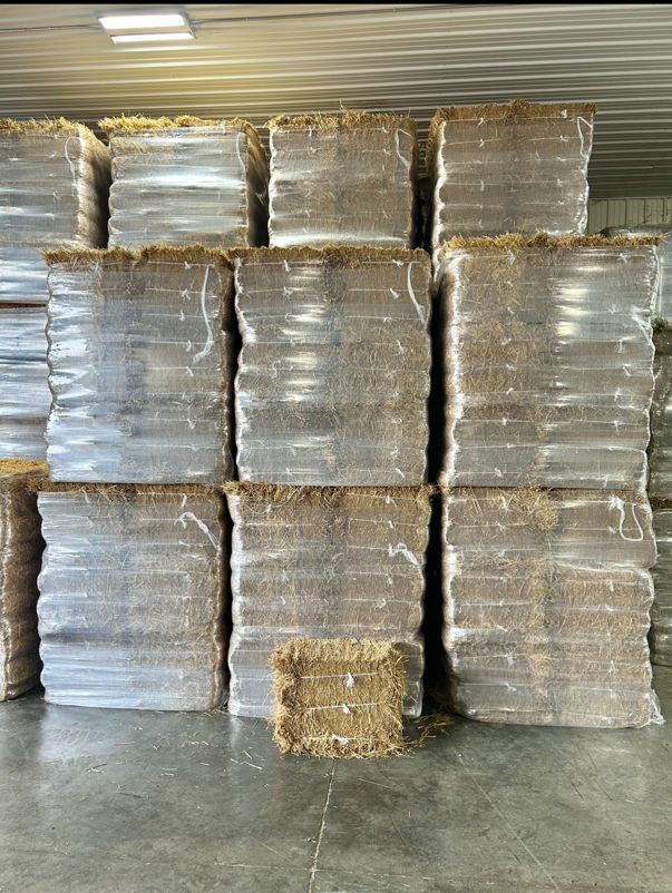 Pallets of wrapped brown compressed material stacked indoors, under white fluorescent lights.