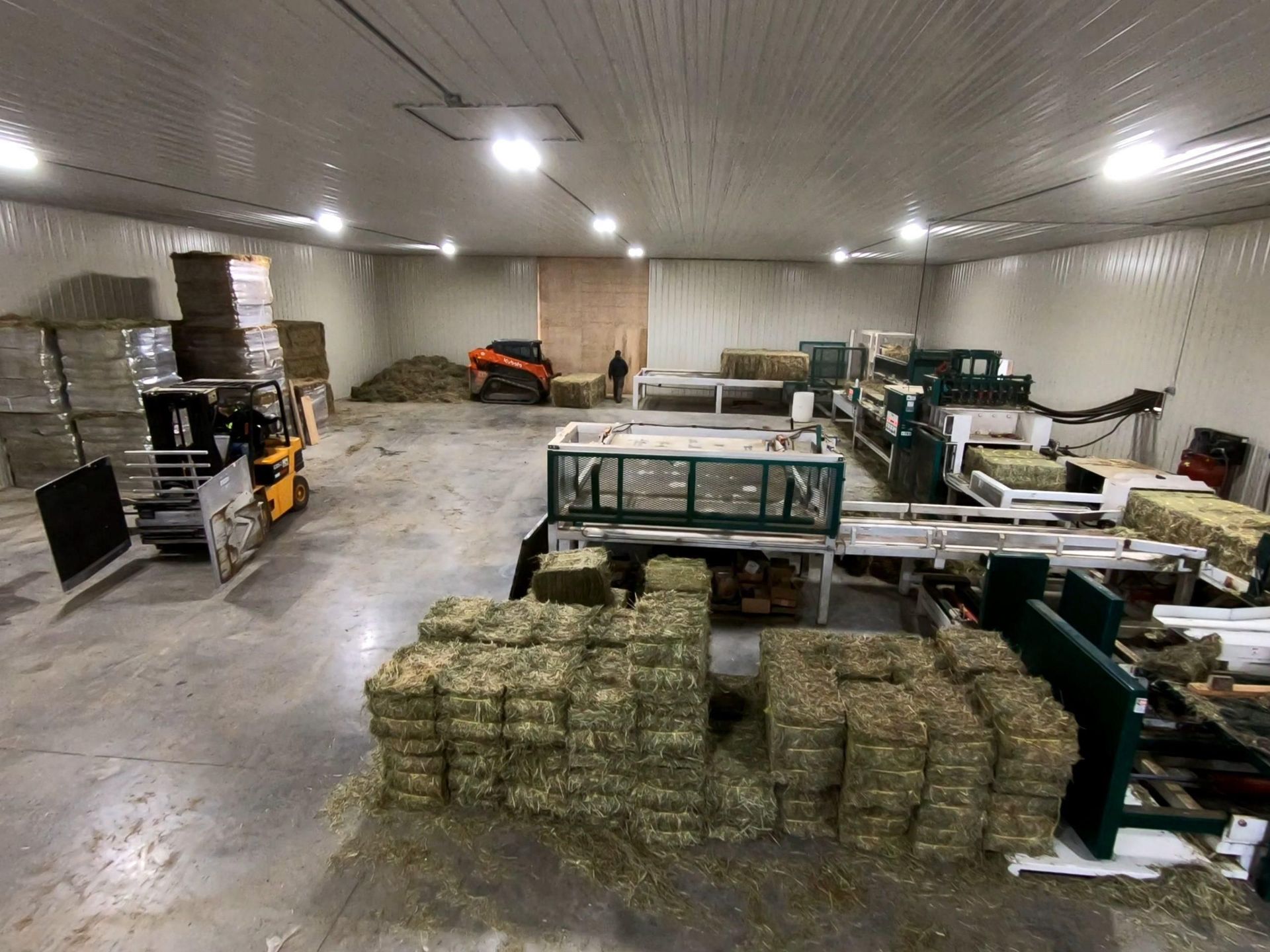 Inside a large warehouse with hay bales and machinery, a person and a small orange tractor.