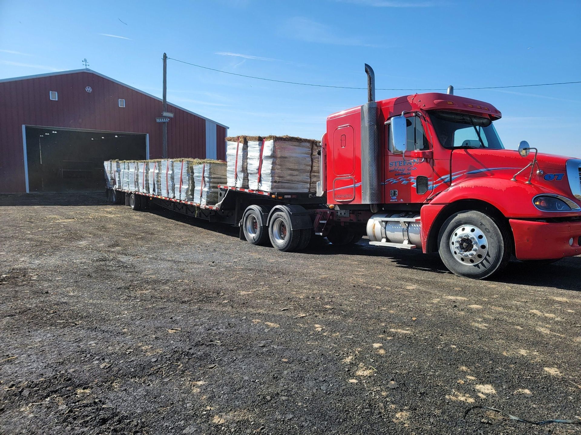Red semi-truck with flatbed trailer loaded with lumber outside a red barn on a sunny day.