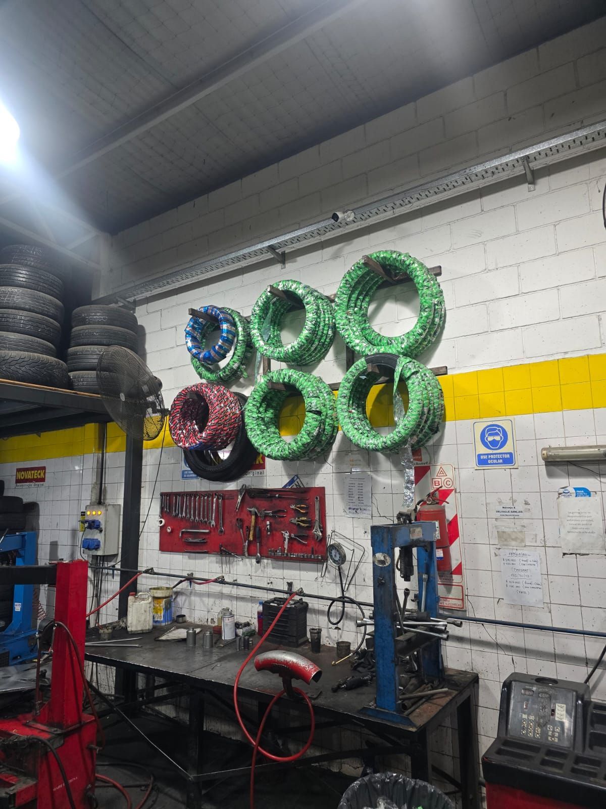 Interior de un taller de reparación de neumáticos con neumáticos apilados, herramientas y mangueras enrolladas colgadas en una pared de ladrillos blancos.