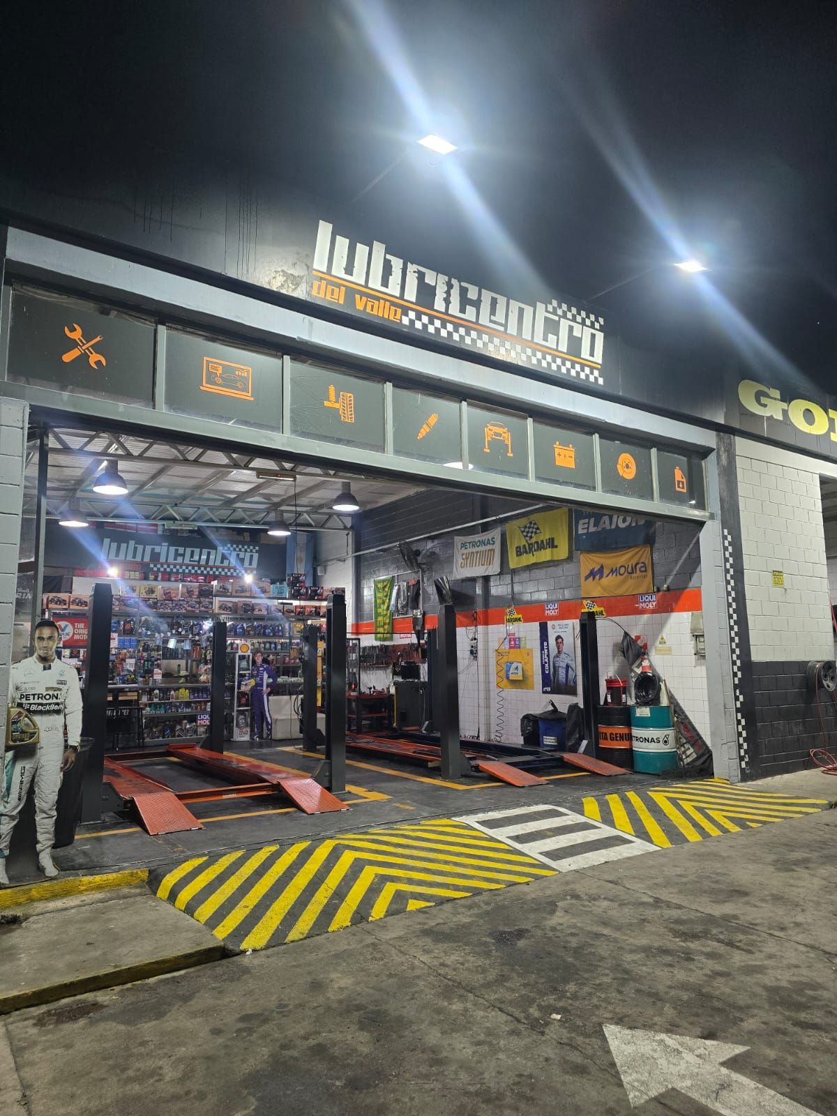 Entrada del taller mecánico Lubricentro, abierta de noche con detalles en naranja y negro. Elevadores de coches visibles.